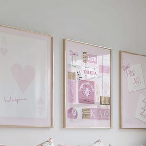 Puede incluir: Tres impresiones enmarcadas con marcos dorados. La impresión de la izquierda es un As de Corazones rosa con el texto "how lucky are we". La impresión central tiene varios gráficos rosas y blancos. La impresión de la derecha tiene dos tarjetas con lazos.