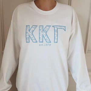 May include: A white sweatshirt with the Greek letters KKT in light blue, floral-patterned embroidery. The text "est. 1870" is below the letters. The sweatshirt has a crew neck and long sleeves.