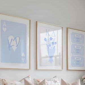 Op de afbeelding: Drie ingelijste kunstdrukken met gouden frames. De prints hebben lichtblauwe ontwerpen op een witte achtergrond. Eén print is een harten aas met "KD" in het midden. Een andere heeft een boeket bloemen. De derde heeft drie tickets.
