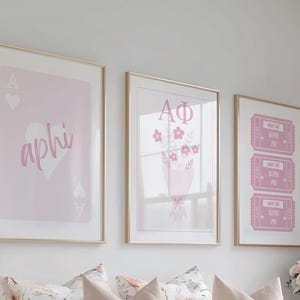 May include: Three framed prints with gold frames. The prints feature pink designs: a playing card with "aphi", a bouquet with "AΦ", and three pink tickets with "Alpha Phi". The prints are displayed above a floral patterned pillow.