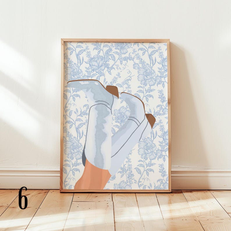 May include: A framed art print featuring a stylized illustration of light blue cowboy boots with brown accents, set against a floral wallpaper background in shades of blue and white. The artwork is displayed on a wooden floor.
