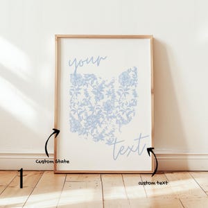 May include: A framed print featuring a floral design in the shape of Ohio, in light blue on a cream background. The words "your" and "text" are written in a script font. The frame is made of light wood.