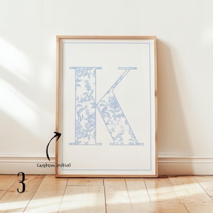 May include: A framed print featuring the letter "K" in a light blue floral pattern. The initial is set against a white background with a light blue border. The frame is made of light-colored wood, and the print is displayed on a wooden floor.