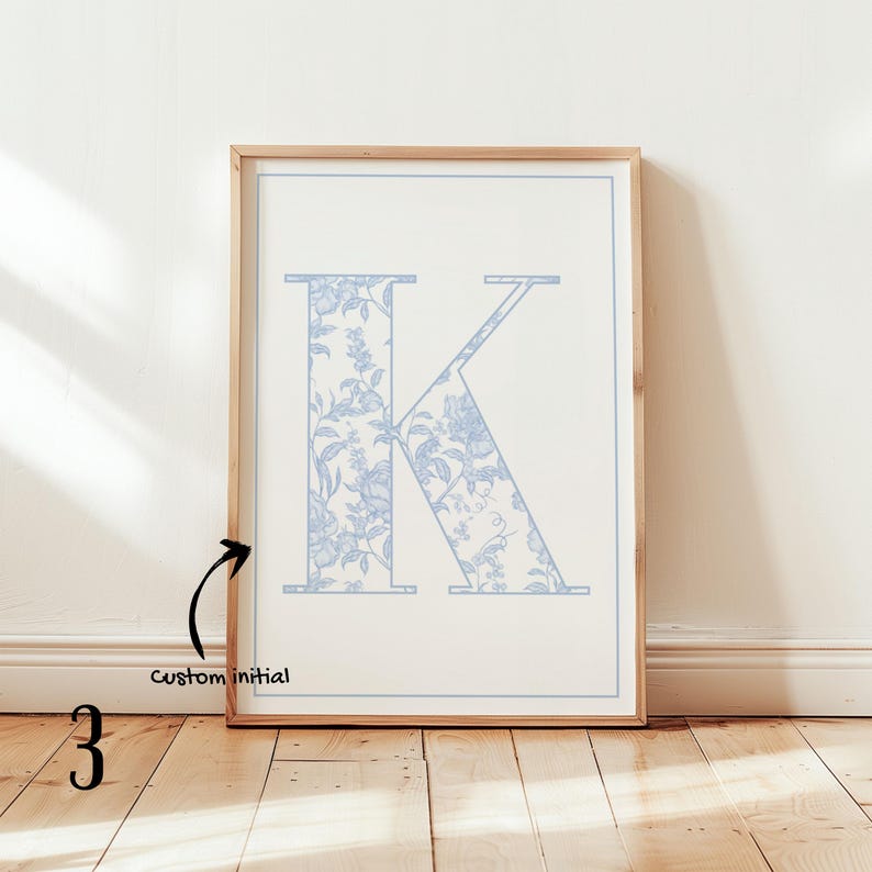 May include: A framed print featuring the letter "K" in a light blue floral pattern. The initial is set against a white background with a light blue border. The frame is made of light-colored wood, and the print is displayed on a wooden floor.