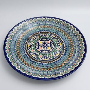 May include: A round ceramic plate featuring a complex, hand-painted geometric and floral pattern in shades of deep blue, turquoise, green, yellow, and white. The design radiates from a central star motif toward the dark blue rim.