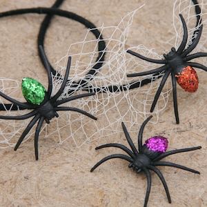 May include: Three black plastic spiders with glitter accents in green, orange, and purple. The spiders have long legs and are positioned near a white web and black string, suggesting a Halloween theme.