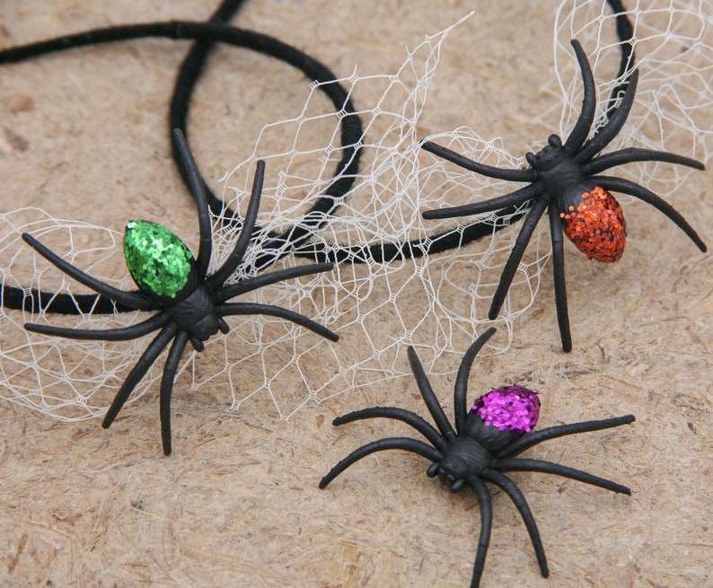 May include: Three black plastic spiders with glitter accents in green, orange, and purple. The spiders have long legs and are positioned near a white web and black string, suggesting a Halloween theme.