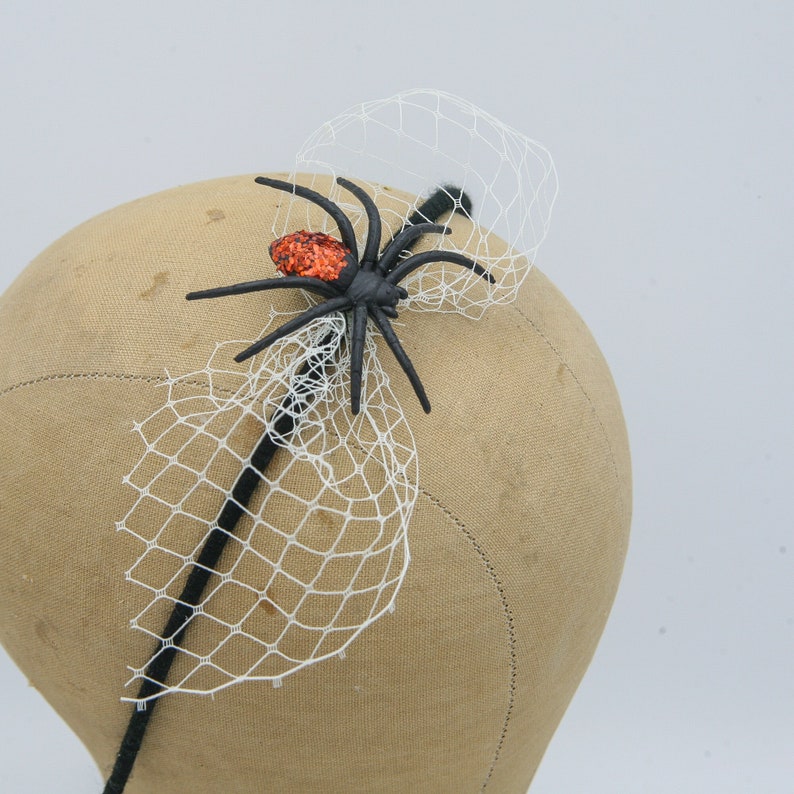 May include: A black spider hair accessory with an orange glitter abdomen and a white net veil. The spider is attached to a black headband, perfect for Halloween or themed events. The accessory is displayed on a mannequin head.