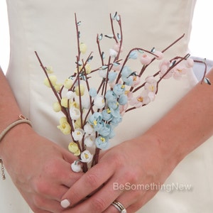 May include: A bouquet of delicate paper flowers in shades of blue, pink, and yellow. The flowers are arranged on thin brown stems and are held in the hands of a person.