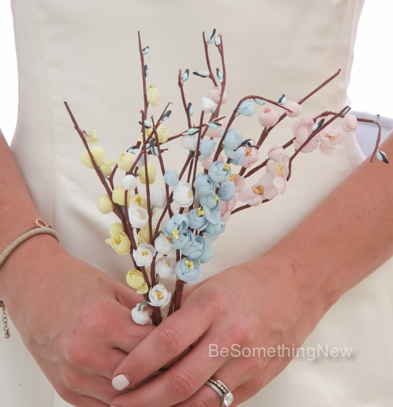 May include: A bouquet of delicate paper flowers in shades of blue, pink, and yellow. The flowers are arranged on thin brown stems and are held in the hands of a person.