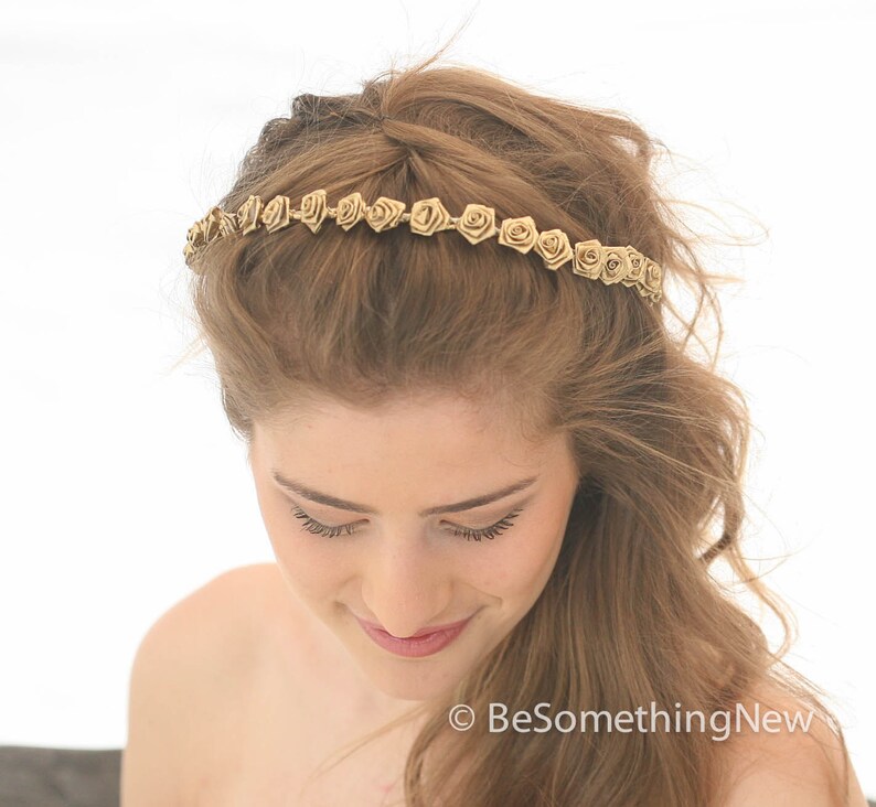 May include: A gold headband with small, delicate fabric rose embellishments. The headband is worn by a person with long, wavy, light brown hair.