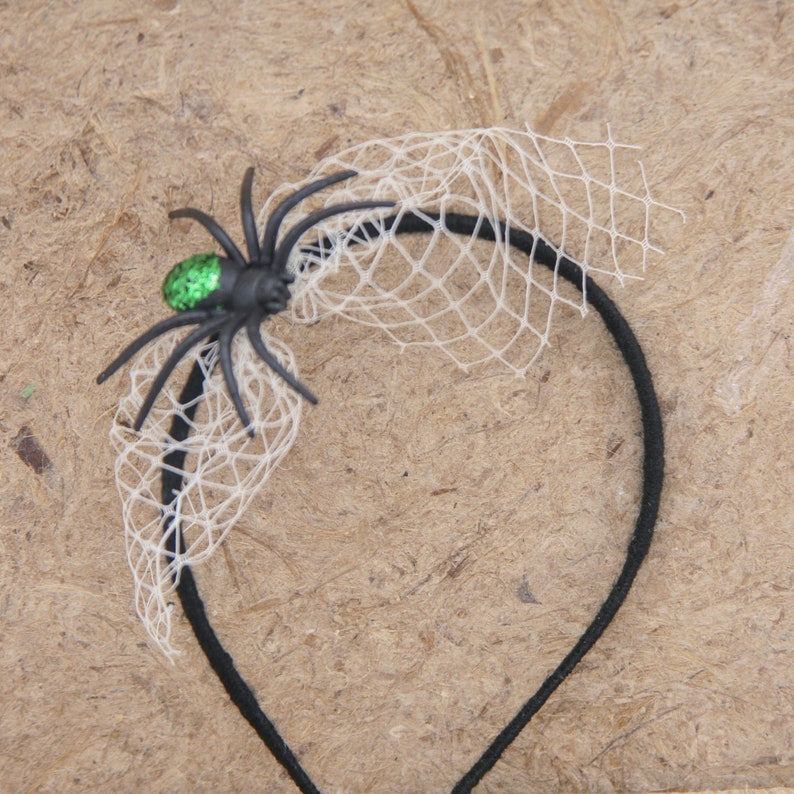 May include: A black headband with a decorative spider and web design. The spider is black with a green glitter abdomen. A white net is attached to the headband, creating a web effect. This Halloween-themed accessory is perfect for costumes.