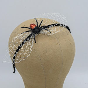 A black spider with orange glitter on its body is attached to a black headband with white tulle netting. The headband is on a brown mannequin head.