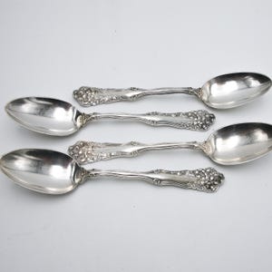May include: Four silver-toned metal spoons with ornate, embossed floral detailing on the handles. Each spoon features a smooth, oval-shaped bowl and a decorative, textured pattern at the base of the handle, showcasing a polished metallic finish.