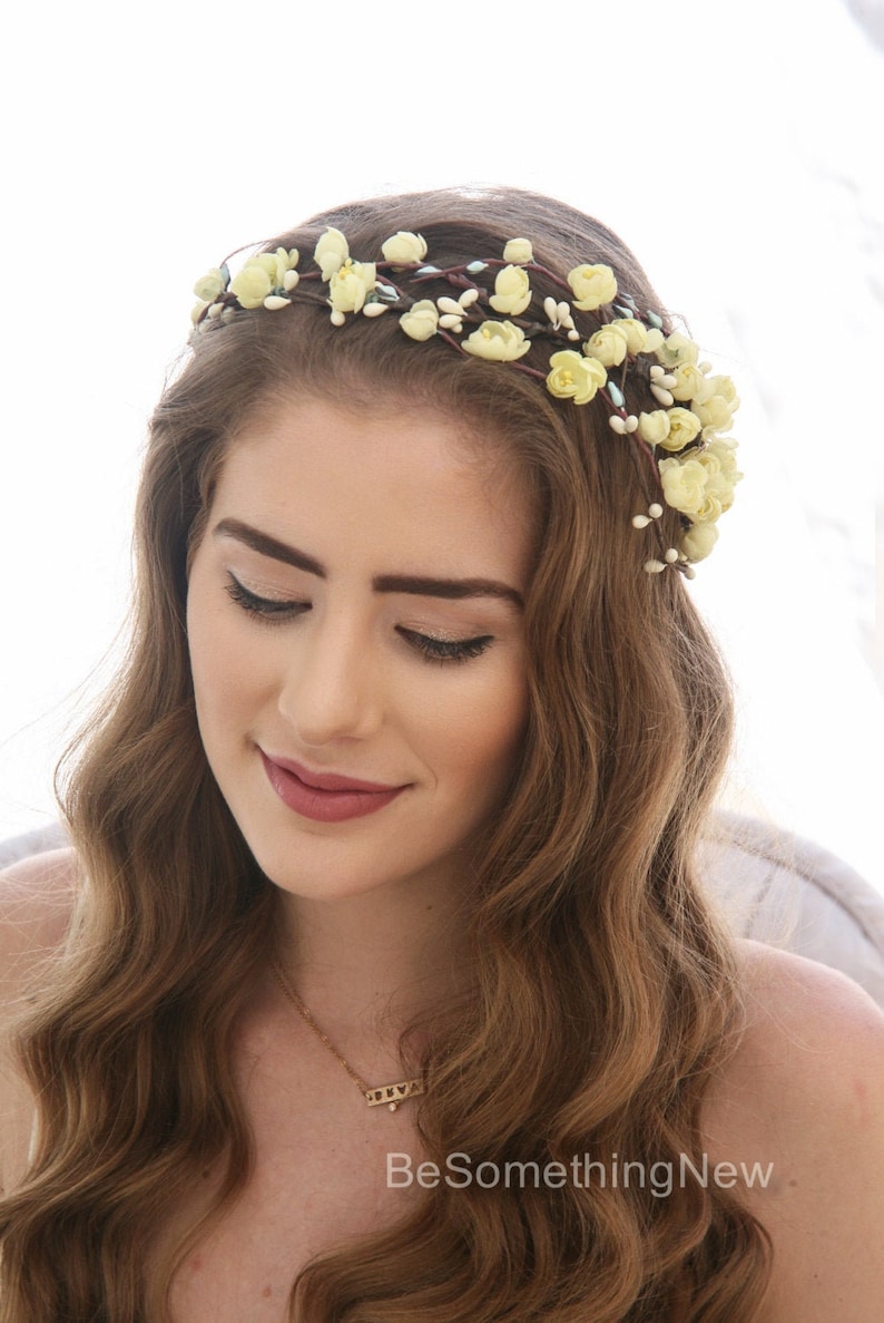 May include: A woman with long brown hair wearing a yellow floral crown. She is wearing a gold necklace with a heart pendant. The necklace has the text "BeSomethingNew" on it.