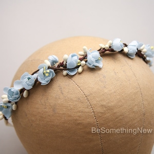 May include: A light blue floral crown with white accents. The crown is made of fabric flowers and is displayed on a brown mannequin head.
