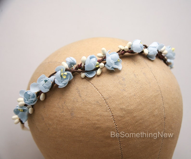 May include: A light blue floral crown with white accents. The crown is made of fabric flowers and is displayed on a brown mannequin head.