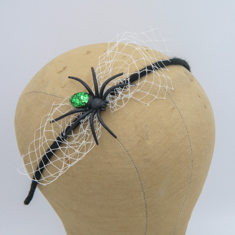 May include: A black headband with a spider and web design. The spider has a green sequined body and is attached to a black headband with a white web overlay. A festive accessory for Halloween.