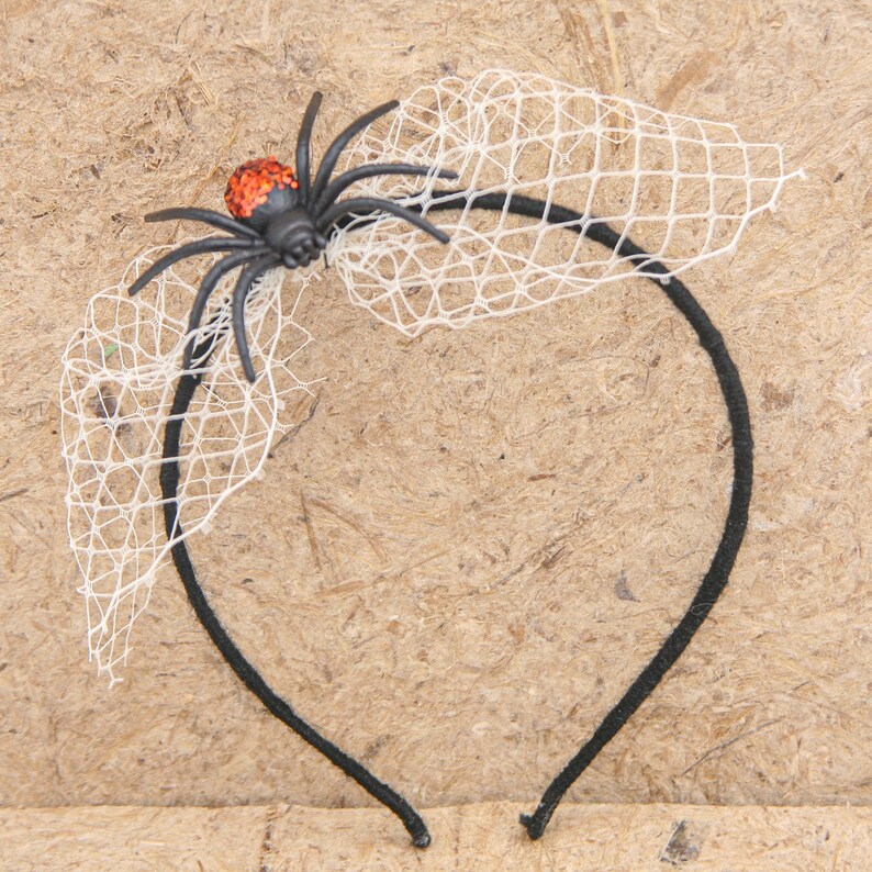 May include: A black headband with a decorative black spider and white netting. The spider has an orange glittery body. The netting is attached to the headband, creating a Halloween-themed accessory.
