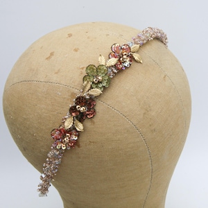 May include: A beaded headband with floral and leaf accents. The headband features a row of small, iridescent beads. The floral elements include green, red, and gold sequins, with gold-coloured metal leaves. The headband is displayed on a mannequin head.