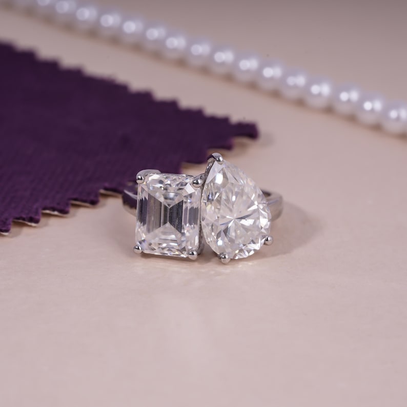 May include: A silver ring with two large, clear gemstones. One is emerald-cut, the other is pear-shaped. The ring is set against a light pink background, with a purple fabric and a string of pearls in the background.