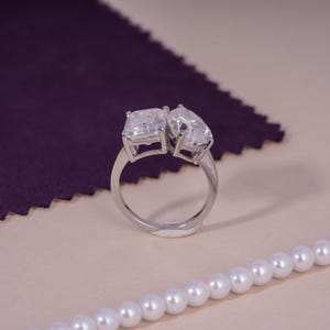 May include: A silver ring featuring two clear, rectangular and pear-shaped stones. The ring is set against a purple fabric and a string of white pearls.
