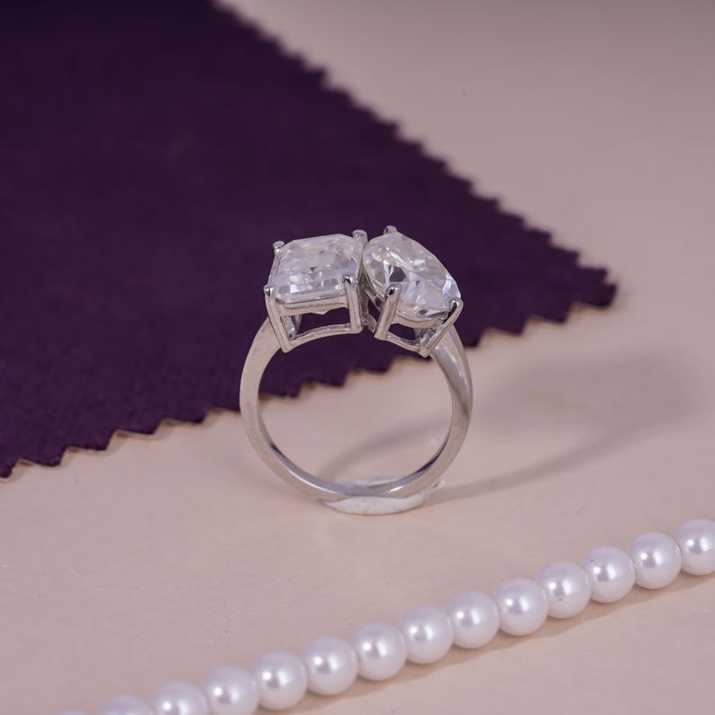 May include: A silver ring featuring two clear, rectangular and pear-shaped stones. The ring is set against a purple fabric and a string of white pearls.