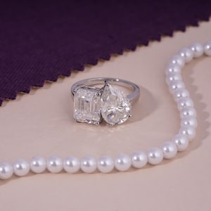 May include: A silver ring with two large, clear gemstones: an emerald cut and a pear-shaped stone. The ring is set against a neutral background, with a strand of white pearls and a purple fabric in the background.