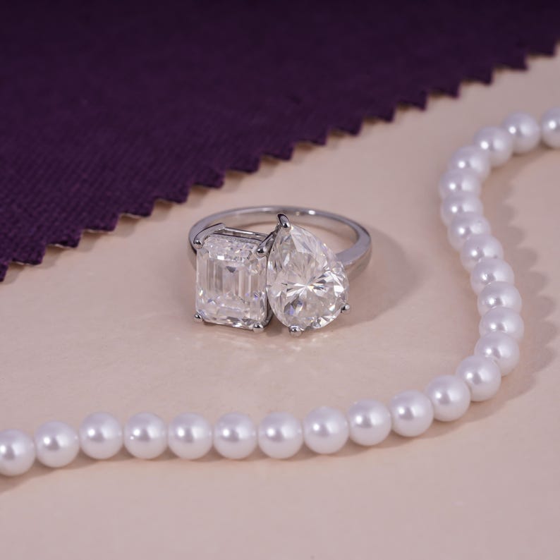 May include: A silver ring with two large, clear gemstones: an emerald cut and a pear-shaped stone. The ring is set against a neutral background, with a strand of white pearls and a purple fabric in the background.