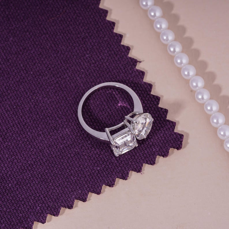 May include: A silver ring with two large, clear gemstones, one square and one heart-shaped, rests on a textured purple fabric. A strand of white pearls is positioned to the right, creating a contrast with the ring's sparkle.