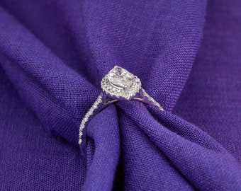 Puede incluir: Un anillo de compromiso de diamante de forma de pera con un engaste halo. El anillo está hecho de oro blanco y presenta una fila de diamantes más pequeños alrededor de la piedra central.