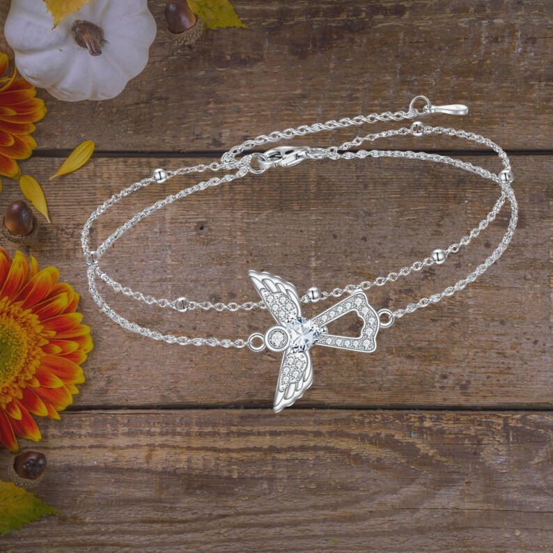 May include: A silver chain anklet with a crystal-encrusted angel charm. The anklet is on a wooden surface with autumnal decorations.