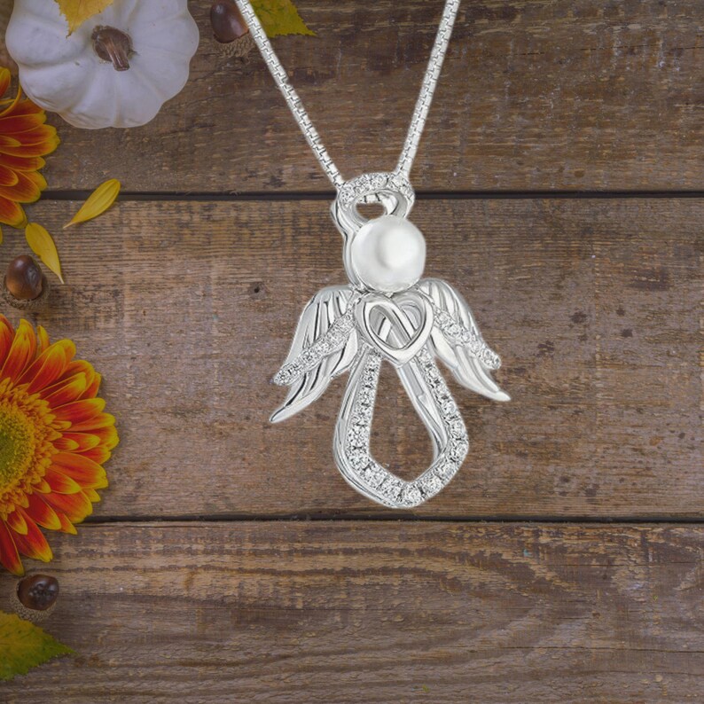 Puede incluir: Un collar con un colgante de &aacute;ngel de plata, con una perla en el centro y un dise&ntilde;o de coraz&oacute;n. El collar est&aacute; sobre un fondo de madera con hojas de oto&ntilde;o.