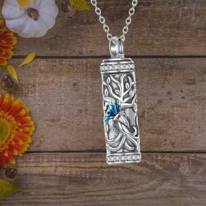 Może przedstawiać: Srebrny naszyjnik z zawieszką w kształcie drzewa życia i niebieskiego motyla. Zawieszka jest urną kremacyjną do przechowywania prochów.