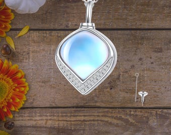 May include: A silver pendant necklace with a teardrop-shaped iridescent stone set in a silver frame. The pendant is surrounded by small clear stones. The necklace is on a wooden surface.