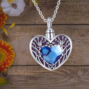 May include: Silver heart-shaped cremation necklace with a blue crystal in the center. The heart is decorated with a tree branch design.