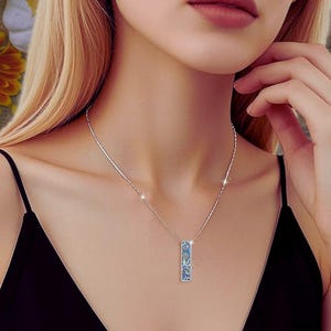 May include: A silver necklace with a rectangular pendant. The pendant has a blue floral design. The necklace is worn by a person with blonde hair and a black top.