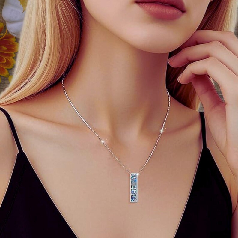 May include: A silver necklace with a rectangular pendant featuring a blue floral design. The pendant hangs from a delicate chain, creating a simple yet elegant look. The necklace is worn against a black top.