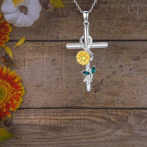 Könnte beinhalten: Silberfarbene Kreuz-Anhänger-Halskette mit Sonnenblume. Das Kreuz hat ein Unendlichkeitszeichen mit dem Schriftzug "You are my sunshine". Der Anhänger hat eine gelbe Sonnenblume, grüne Blätter und klare Edelsteine. Die Kette ist an einer silbernen Kette.