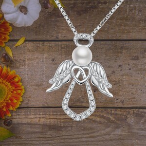 Puede incluir: Un collar con un colgante de &aacute;ngel de plata con una perla y detalles de cristal transparente. El colgante presenta un dise&ntilde;o de coraz&oacute;n y alas de &aacute;ngel.