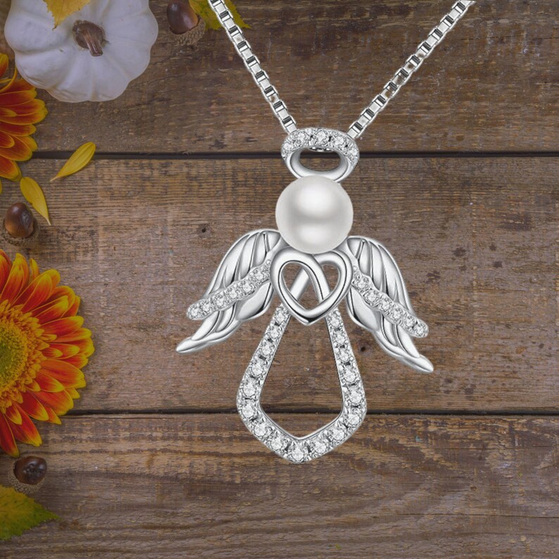 Puede incluir: Un collar con un colgante de &aacute;ngel de plata con una perla y detalles de cristal transparente. El colgante presenta un dise&ntilde;o de coraz&oacute;n y alas de &aacute;ngel.