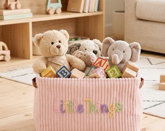 Könnte beinhalten: Ein rechteckiger Aufbewahrungskorb aus rosa Kord mit braunen Seitengriffen und dem gestickten Text Little Things in Pastellfarben. Der Korb ist mit einem beigen Teddybären, einem grauen Elefanten-Plüschtier und mehreren hölzernen Buchstabenwürfeln gefüllt.