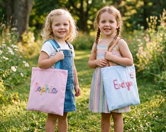 May include: Two rectangular fabric tote bags, one light pink with the name Sophia in colorful letters and one light blue with the name Evelyn in pink letters, each featuring two shoulder straps and a small flower embellishment on the blue bag.