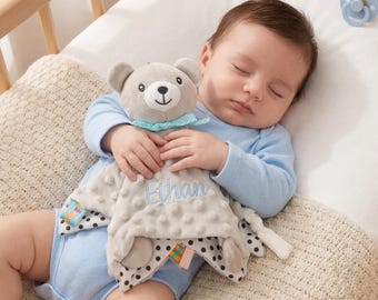 Könnte beinhalten: Ein schlafendes Baby in einem hellblauen Strampler kuschelt mit einer grauen Teddybär-Kuscheldecke. Die Decke hat ein gepunktetes Muster und den Namen "Ethan" in Blau gestickt. Ein Schnuller und ein Beißring sind sichtbar.