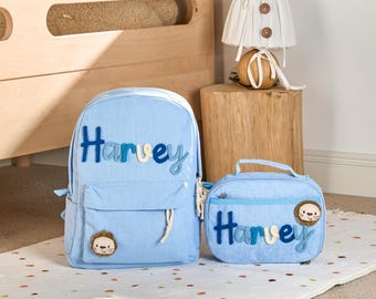Könnte beinhalten: Ein hellblaues Rucksack- und Lunchpaket-Set mit vertikalen Streifen. Der Name "Harvey" ist in blauen und weißen Buchstaben auf beiden Artikeln gestickt. Jeder Artikel hat einen Löwengesicht-Aufnäher. Das Set ist für Kinder konzipiert.