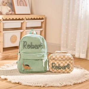 May include: A sage green corduroy backpack and a matching beige and white checkered lunch bag. Both items feature the name Robert embroidered in dark green script. The backpack includes a small orange dinosaur patch on the front pocket.