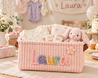 Könnte beinhalten: Ein rechteckiger Aufbewahrungskorb aus rosa Kord mit dem Namen Laura in mehrfarbigen Strickbuchstaben und einer kleinen Blumenapplikation. Der Korb enthält einen weißen Baby-Body, ein gemustertes Mulltuch, Babysocken und einen rosa Plüschhasen.