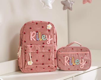 Könnte beinhalten: Ein rosa Cord-Rucksack und eine passende Lunchtasche, beide mit einem Erdbeermuster und dem Namen "Riley" in bunten Buchstaben bestickt. Der Rucksack hat eine Vordertasche und ein Blumenmuster.