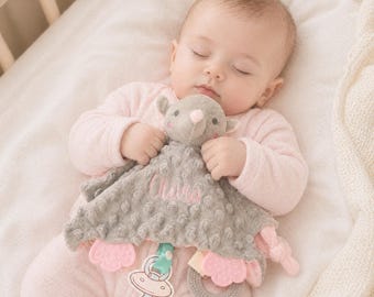 Könnte beinhalten: Eine graue und rosa Baby-Sicherheitsdecke mit einem Plüsch-Mauskopf und dem Namen "Olivia" in Rosa gestickt. Die Decke hat eine strukturierte Oberfläche und einen befestigten Beißring und Schnullerclip. Das Baby trägt ein rosa Outfit.