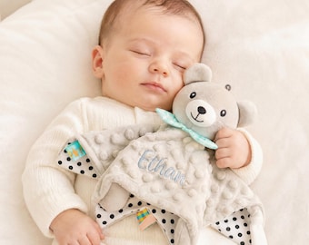 Könnte beinhalten: Ein schlafendes Baby in einem weißen Strampler, das eine graue Bär-Kuscheldecke hält. Die Decke hat ein gepunktetes Muster, den Namen "Ethan" gestickt und eine hellblaue Schleife.